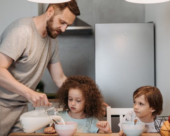 A father pours milk into cereal bowls for his children at the breakfast table in a modern kitchen.